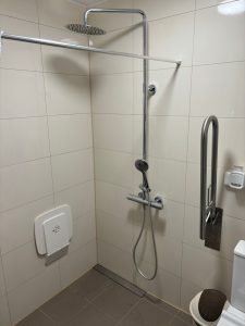 Dušas zona ar bēšām flīzēm, aprīkota ar lietus dušu, rokas dušu, saliekamu sēdekli un atbalsta rokturi, kas nodrošina pieejamību.