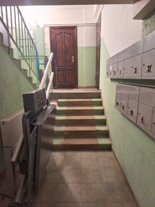 Pieejama dušas zona ar lietus dušu, rokas dušu, pie sienas stiprinātu saliekamu sēdekli un atbalsta rokturiem uz gaišām flīzētām sienām un pelēkas grīdas.