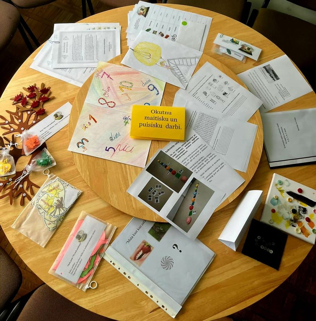 Apaļš koka galds, kas klāts ar dažādiem mācību un radošajiem materiāliem: drukātām darba lapām, bērnu krāsainiem zīmējumiem ar cipariem un figūrām, kā arī rokdarbiem, piemēram, koka koku ar sarkaniem lapu rotājumiem un maisiņiem ar pērlēm. Galda centrā ir dzeltens bloks ar tekstu: "Okūtnu maitisku un puisisku darbi." Redzamas arī lapas ar fotogrāfijām, mazas dekorētas audekla plāksnes ar pogām un krāsu, un lapa ar zemeņu un jautājuma zīmes attēlu.