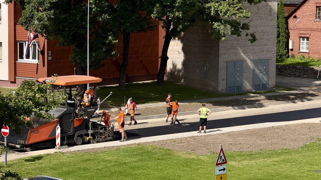 Celtniecības strādnieki oranžos un dzeltenos tērpos klāj jaunu asfaltu uz ielas ar oranžu un melnu asfaltbetona ieklājēju Volvo P6820D. Daži strādnieki manuāli apstrādā asfaltu. Fonā redzamas ēkas, tostarp viena ar Latvijas karogu, un koki, priekšplānā – ceļa darbu zīme.