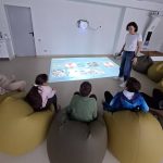 Cilvēki veido smiltis interaktīvā smilšu kastē, kurā uz reljefa tiek projicētas zilas (ūdens) un zaļas (zeme) krāsas, zilā apgaismojumā.