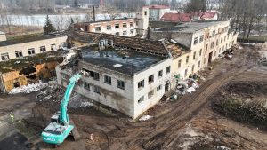 Liels tirkīza krāsas "Kobelco" ekskavators nojauc baltu ķieģeļu ēku vecu, nolaistu ēku kompleksā. Apkārt ir gruveši un dubļaina zeme, fonā redzams aizsalis ūdens un kaili koki. Viens strādnieks dzeltenā vestē stāv blakus ekskavatoram.
