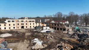 Plaša nojaukšanas vieta ar veciem, daļēji nojauktiem namiem. Pa kreisi redzama liela dzeltenīga ēka ar tukšām logu ailēm, kurai blakus strādā oranžs ekskavators. Pa labi ir ķieģeļu ēkas drupas, pie kurām darbojas tirkīzzils ekskavators. Zeme ir klāta ar dubļiem, gruvešiem un sniega paliekām, zem skaidrām zilām debesīm.