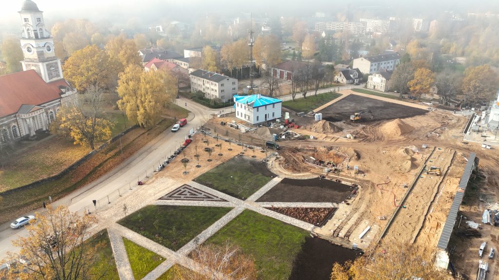 Augšupējs skats uz pilsētas laukumu rudens dienā, kur norisinās apjomīgi rekonstrukcijas darbi. Laukuma daļā ir ieklāts zaļš zālājs un bruģēti celiņi ar ģeometriskiem rakstiem, bet lielāko daļu veido būvlaukums ar smilšu kaudzēm un celtniecības tehniku. Blakus redzama balta ēka ar zilu jumta pārklāju un veca baznīca ar sarkanu jumtu un baltu torni, ko ieskauj dzelteni rudenīgi koki.