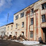 Vecas, nolaistas dzeltenīgi-sarkanas ēkas fasāde ar nolobītu apmetumu, redzamiem ķieģeļiem un logiem. Pie tās pamatnes ir koka būvgružu kaudzes uz sniegotas un dubļainas zemes. Kreisajā pusē redzams funkcionējošs industriāls komplekss ar augstu, dūmojošu skursteni zem ziliem debesīm.