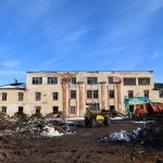 Liela, nolaista daudzstāvu ēka ar bojātu fasādi un izsistiem logiem. Tās priekšā ir dubļains būvlaukums ar būvgružu kaudzēm, dzeltenu pašizgāzēju un tirkīzzilu KOBELCO ekskavatoru pie oranžas kravas automašīnas. Skaidras zilas debesis.