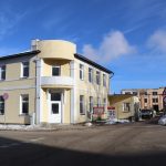 Dzeltenīgi krāsota divstāvu ēka ar noapaļotu stūri un balkonu. Priekšplānā sniega kaudzes un ceļš, kas ved uz vecākām, nolietotām ēkām. Redzams brīdinājuma plakāts ar uzrakstu "IEJA AIZLIEGTA" un "BĪSTAMI! STRĀDĀ BŪVTEHNIKA", kā arī ceļa zīmes, kas norāda uz remontdarbiem un iebraukšanas aizliegumu. Zilas debesis ar mākoņiem.