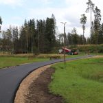 Jauns asfaltēts celiņš līkumo cauri zaļam zālājam. Fonā redzama asfalta ieklāšanas mašīna, kravas auto un strādnieki oranžās vestēs, veicot ceļa būves darbus. Apkārtnē ir mežs, ielas apgaismojuma stabs un tālumā dzeltena ēka.