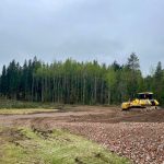 Dzeltenais buldozers D7E stāv būvlaukumā ar rakto zemi un gružu kaudzēm priekšplānā, fonā – mežs zem apmākušām debesīm.