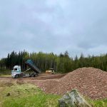 Dzeltens buldozers ar uzrakstu "875E" uz sāniem atrodas attīrītā būvlaukumā. Zeme ir tumši brūna ar riepu sliedēm un sarkanu gružu kaudzi priekšplānā. Fonā redzams blīvs mežs ar zaļiem kokiem zem apmākušām debesīm.