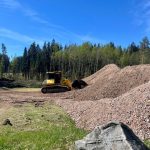 Dzeltenais buldozers atrodas iztīrītā zemes gabalā, iespējams, izlīdzina to, blakus būvgružu kaudzei. Fonā blīvs mežs zem mākoņainām debesīm.