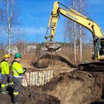 Dzeltenais KOMATSU ekskavators "PC 210 LC" rok zemi būvlaukumā. Trīs strādnieki zaļās un dzeltenās atstarojošās drēbēs un ķiverēs stāv pie izrakta grāvja ar bērzu baļķu nostiprinājumu, fonā koki un mājas.