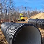 Dzeltena Komatsu ekskavators ar uzrakstiem "KOMATSU" un "PC 210 LC" rok zemes uzbērumu būvlaukumā. Trīs strādnieki spilgtās vestēs un aizsargķiverēs uzrauga darbu. Divi no viņiem stāv pie ūdenstilpes un guļbaļķu atbalsta sienas, bet trešais atrodas izraktā tranšejā. Ekskavatora operators sēž kabīnē. Fonā kaili koki un mājas zem zilas debess.