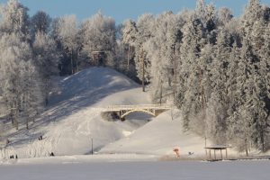 Sniegota ziemas ainava ar aizsalušu ezeru priekšplānā un nelielu koka piestātni labajā pusē. Sniegoti pauguri, kas klāti ar sarmotiem kokiem, ar lapeni uz kreisā paugura virsotnes un baltu arku tiltu starp pauguriem. Vairāki cilvēki slidinās ar ragavām pa sniegoto nogāzi.