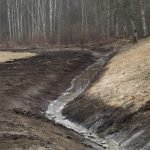 Dzeltenais ekskavators "KOMATSU PC 210 LC" veic rakšanas darbus ar kausu virs zemes kaudzes. Blakus stāv trīs strādnieki aizsargtērpos: divi pie ūdenskrātuves, kuras krasts stiprināts ar koka baļķiem, un viens atrodas tranšejā. Ekskavatora operators redzams kabīnē. Fons ar kailiem kokiem un attālām mājām zem skaidrām zilām debesīm.