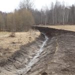 Būvlaukumā pie ūdenstilpes dzeltens ekskavators Komatsu PC 210 LC rok zemi, bet trīs strādnieki spilgtās vestēs un aizsargķiverēs uzrauga darbu. Blakus tranšejai izbūvēta bērza baļķu siena. Fonā kaili koki un mājas zem zilas debess.