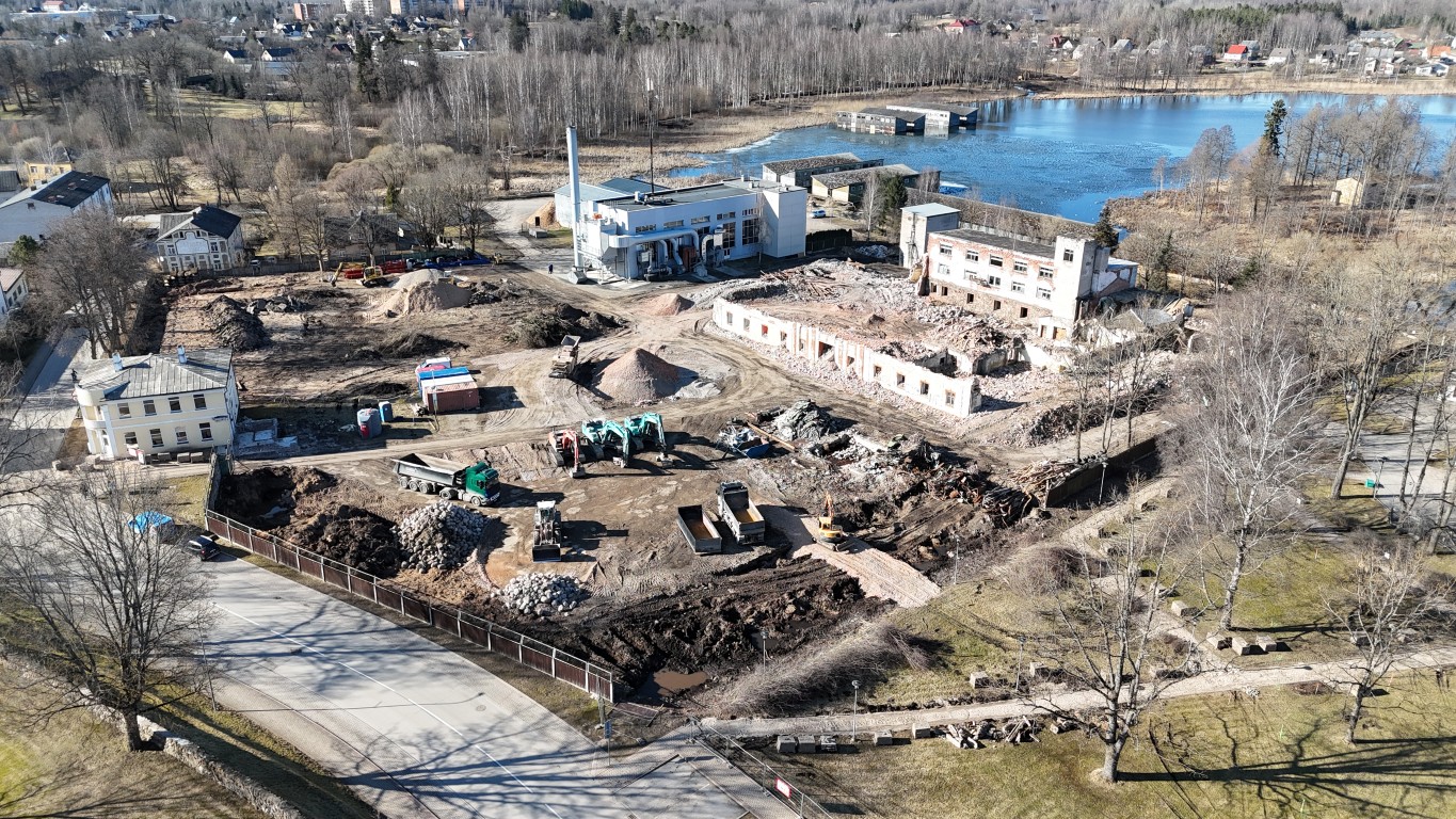 Drona skats uz plašu būvlaukumu pie ūdenskrātuves ar daļēji nojauktām ēkām, kaudzēm drupu un būvniecības tehniku. Blakus atrodas modernas celtnes un daļēji aizsalis ezers, ko ieskauj kaili koki un attālas apbūves silueti.