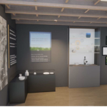 Iekšskats modernā izstāžu telpā ar tumši pelēkām sienām un koka griestiem. Kreisajā sienā liels reljefs topogrāfiskās kartes attēls ar uzrakstu "KĀ RĪJAS MAKI". Blakus tam vertikāls melns panelis ar baltu tekstu. Centrālais ekrāns rāda tekstu "šķiet labāk saprasts dabā" un atkārtotu vārdu "sakot", fonā izplūdis ainavas attēls. Zem ekrāna melna virsma ar baltu bļodu, pudeli un maziem traukiem. Labajā pusē balta stikla vitrīna ar bļodām un glāzēm, zem tās vīna pudele un glāze. Pie sienas septiņi melni rāmji, no kuriem sešos ir ainavu fotogrāfijas un viens ir tukšs.