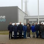 Pie ēkas stāv vienpadsmit cilvēki un divas mašīnas