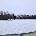 Plašs āra ledus laukums ar saskrāpētu ledu un vieniem hokeja vārtiem kreisajā pusē. Fonā mežs un apmākusies ziemas debess.