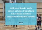 Basketbola laukums ar spēlētājiem, virs kura ir uzklāts zils pārklājums ar tekstu: "Alūksnes Sporta skola saņem Latvijas Basketbola Savienības atbalstu basketbola attīstībai novadā". Redzams arī logo "Alūksne".