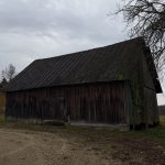 Liela, veca koka klēts ar tumšu, viļņotu jumtu un sūnu vienā sānā. Tai blakus atrodas gaiša ķieģeļu ēka ar koka apšuvumu un sarkanu jumtu, kurai pie sāniem ir sarkana caurule. Pa labi redzams liels kails koks un priekšplānā zemes celiņš zem apmākušām debesīm.