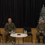 Četri vīrieši uz skatuves ar melniem aizkariem un trim dekoratīvām Ziemassvētku eglītēm. Divi vīrieši kamuflāžas uniformās sēž pie galda ar ūdens pudelēm un mikrofoniem. Viens vīrietis pelēkā žaketē stāv kreisajā pusē, bet otrs vīrietis tumšā džemperī stāv labajā pusē, abiem rokas saliktas.