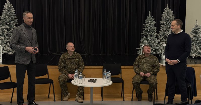 Četri vīrieši uz skatuves ar melnu aizkaru fonu un baltām Ziemassvētku eglītēm. Divi vīrieši civilā apģērbā stāv kreisajā un labajā pusē, bet divi vīrieši kamuflāžas uniformās sēž vidū. Priekšplānā starp sēdošajiem vīriešiem ir galds ar ūdens pudelēm un mikrofoniem.