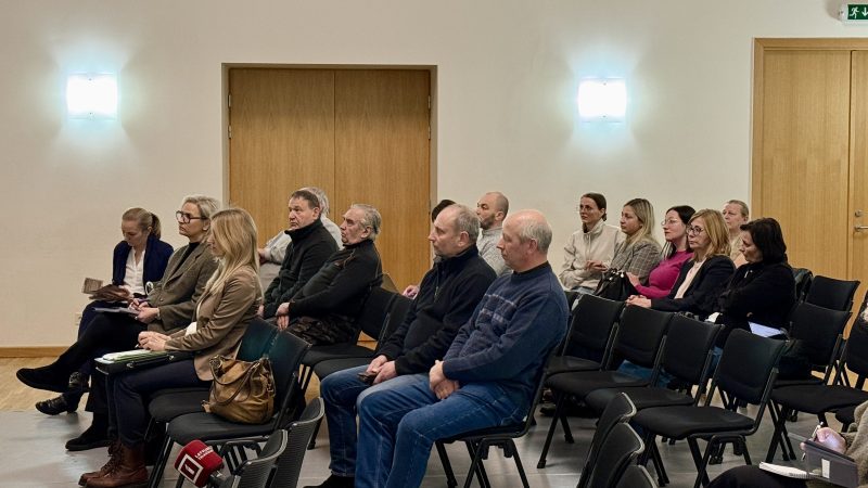 Četri vīrieši uz skatuves ar melniem aizkariem un trim dekoratīvām Ziemassvētku eglītēm. Divi vīrieši kamuflāžas uniformās sēž pie galda ar ūdens pudelēm un mikrofoniem. Viens vīrietis pelēkā žaketē stāv kreisajā pusē, bet otrs vīrietis tumšā džemperī stāv labajā pusē, abiem rokas saliktas.
