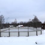 Sniegots āra hokeja laukums vai slidotava ar baltiem bortiem un tīklu, blakus tam atrodas neliela sarkana koka ēka. Apkārtne ir klāta ar sniegu, fonā redzami koki, dažas mājas un pelēkas debesis.