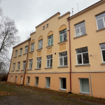 Daudzstāvu ēka, krāsota gaišdzeltenā un oranžā krāsā, stāv zem apmākušām debesīm. Zeme priekšā klāta ar kritušām rudens lapām. Ēkai ir daudz logu ar baltiem rāmjiem, dažiem no tiem ir arkveida virsmas, un dekoratīvi elementi uz jumta malas. Kreisajā pusē redzami kaili koki.