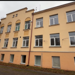 Divstāvu ēka ar gaiši dzeltenu augšējo un oranžu apakšējo fasādi, daudziem baltiem logiem un sarkaniem notekcaurulēm. Priekšplānā bruģēts laukums ar kritušām rudens lapām zem pelēkām debesīm.