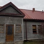 Tumšs koka šķūnis ar sūnainu jumtu un koka durvīm priekšplānā. Kreisajā pusē redzama gaišāka ēka ar baltiem ķieģeļiem un sarkanu jumtu, bet labajā pusē stāv kails koks. Diena ir apmākusies.