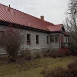 Veca koka ēka ar tumšu, rievotu jumtu, daļēji apaugušu ar sūnām, atrodas grants ceļa malā. Labajā pusē redzams liels kails koks, bet fonā – gaišu ķieģeļu māja ar sarkanu jumtu zem apmākušām debesīm.