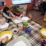 Pieaugušo grupa apmeklē prezentāciju gaišā telpā. Sieviete stāv priekšā, rādot uz projektora ekrānu, kurā redzams "eTwinning" un "2023./2024.m.g." kopā ar grupu attēliem. Citi dalībnieki sēž ar piezīmju grāmatiņām, vērsti pret prezentētāju. Priekšplānā ir galds ar tēju un uzkodām.