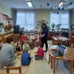 Cilvēku grupa gaišā telpā apmeklē prezentāciju. Sieviete stāv pie ekrāna, kurā redzams slaids ar tekstu "eTwinning" un "2023.2024.m.g" un attēliem no aktivitātēm. Klātesošie sēž, daži ar piezīmju grāmatiņām, un priekšplānā ir galds ar atspirdzinājumiem.