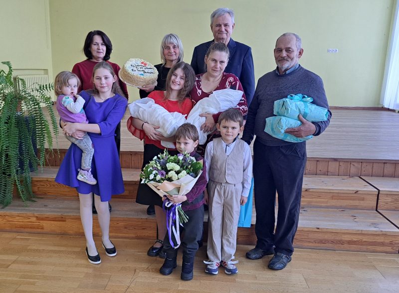 Deviņi ģimenes locekļi – bērni, pieaugušie un seniori – pozē svinību foto. Sieviete tur torti ar uzrakstu "Daudz laimes dzimšanas dienā! Es tevi mīlu.". Cita sieviete rokās tur jaundzimušu zīdaini. Jaunāka meitene tur mazu bērnu, zēns – ziedu pušķi, un vecāks vīrietis – divas iesaiņotas dāvanas.
