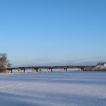 Garš, zems gājēju tilts stiepjas pāri sniegotam, aizsalušam ezeram vai līcim. Uz tilta staigā daži cilvēki. Fonā redzami koki un skaidras zilas debesis.