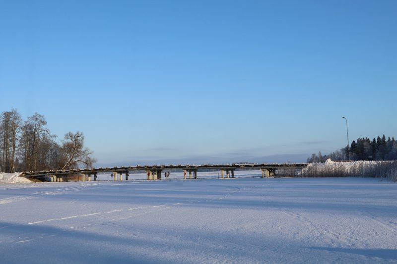 Garš, zems gājēju tilts stiepjas pāri sniegotam, aizsalušam ezeram vai līcim. Uz tilta staigā daži cilvēki. Fonā redzami koki un skaidras zilas debesis.