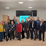 Cilvēku grupa, kas sastāv gan no sievietēm, gan vīriešiem, pozē fotogrāfijai modernā telpā. Fonā redzams liels ekrāns ar uzrakstu "Alūksne" un Latvijas un Eiropas Savienības karogi.