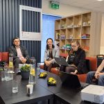 Pieci cilvēki (četras sievietes un viens vīrietis) sēž uz oranža dīvāna, skatoties pa labi. Aiz viņiem uz sienas lieliem burtiem rakstīts "PA GALMS". Trīs sievietes kreisajā pusē valkā melnus t-kreklus ar uzrakstiem "PA GALMS" un "ALŪKSNES BĒRNU UN JAUNIEŠU CENTRS". Priekšplānā uz galda ir klēpjdators, dzērieni un uzkodas. Sieviete labajā malā tur krūzi ar redzamu uzrakstu "LAI SĀKĀS".