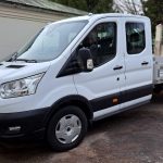 Balts Ford Transit furgons ar dubulto kabīni un kravas platformu, reģistrācijas numurs LV-9812, novietots ārpus telpām uz grants seguma.