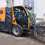 Oranži melns ielu tīrīšanas transportlīdzeklis ar uzrakstiem "SX2", "HOLDER", "ALWARK", "muvo" un "FIRASCO" ir novietots uz asfalta pie vecas ēkas ar gaišām sienām un logiem. Transportlīdzekļa priekšpusē ir piestiprinātas lielas rotējošas slaucīšanas birstes.