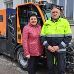 Sieviete sarkanā jakā un vīrietis dzeltenā augstas redzamības darba jakā stāv blakus oranžai ielu tīrīšanas mašīnai. Uz mašīnas ir redzami zīmoli "ALWARK INDUSTRIAL CLEANING", "R Rosco" un "muvo". Fonā gaiša ēka.