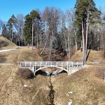 Agrs pavasaris parkā, kur balts koka tilts šķērso nelielu strautu starp brūniem, sausiem pauguriem ar kūstoša sniega paliekām. Uz attālāka pakalna redzams apaļš akmens templis, apkārt kaili lapu koki un zaļas priedes zem skaidrām zilām debesīm.