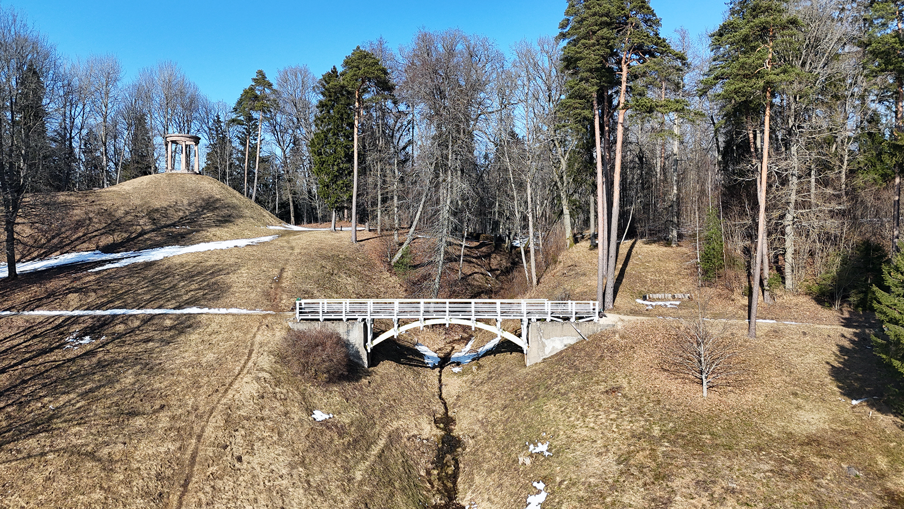 Agrs pavasaris parkā, kur balts koka tilts šķērso nelielu strautu starp brūniem, sausiem pauguriem ar kūstoša sniega paliekām. Uz attālāka pakalna redzams apaļš akmens templis, apkārt kaili lapu koki un zaļas priedes zem skaidrām zilām debesīm.