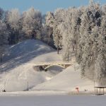 Sniegota ziemas ainava ar sasalušu ezeru, sniegotām nogāzēm, kur vizinās cilvēki, baltu arku tiltu un koka lapeni uz apsarmojušu koku klāta kalna.