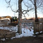 Dzeltens CAT 315F ekskavators stāv sniegotā un niedru aizaugušā vietā. Uz bērza stumbra redzams sarkans marķējums "17", bet zemē iesprausti sarkani stabiņi. Attēla labajā pusē redzama koka sēta. Fonā, zem zilu debesu fona, ir redzamas ēkas un baznīcas tornis.