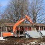 Oranžs Hitachi 210 ekskavators ar demontāžas satvērēju nojauc daļēji nojauktu sarkanbaltu ēku ar redzamu jumta karkasu. Fonā redzami kaili koki zem zilām debesīm, priekšplānā sniegs un būvgruži.