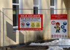 Pie metāla žoga pie dzeltenas ēkas ar logiem, kurā notiek būvdarbi, piestiprinātas divas informatīvas zīmes. Kreisajā sarkanajā zīmē teikts "IEEJA AIZLIEGTA ŠEIT NOTIEK BŪVDARBI: OBJEKTĀ JĀIEVĒRO DARBA AIZSARDZĪBAS, UGUNSDROŠĪBAS UN VIDES AIZSARDZĪBAS PRASĪBAS! ĀRKĀRTĒJĀ SITUĀCIJĀ ZVANĪT 112" ar ikonām par ieejas aizliegumu, obligātu ķiveres lietošanu, vispārēju bīstamību un paceltu kravu. Labajā zīmē, kas ir balta ar sarkanu virsrakstu, rakstīts "UZMANĪBU! KRAVAS AUTOMOBIĻU VADĪTĀJIEM ATRODOTIES ĀRPUS TRANSPORTLĪDZEKĻA JĀLIETO: ATTENTION! WHEN OUTSIDE THE VEHICLE TRUCK DRIVERS MUST WEAR:", ar piktogrammu, kas attēlo celtnieku un uzskaita obligāto aizsargaprīkojumu (ķivere, veste, zābaki), ātruma ierobežojumu 5 km/h, un citas drošības zīmes. Priekšplānā uz zemes ir sniegs.